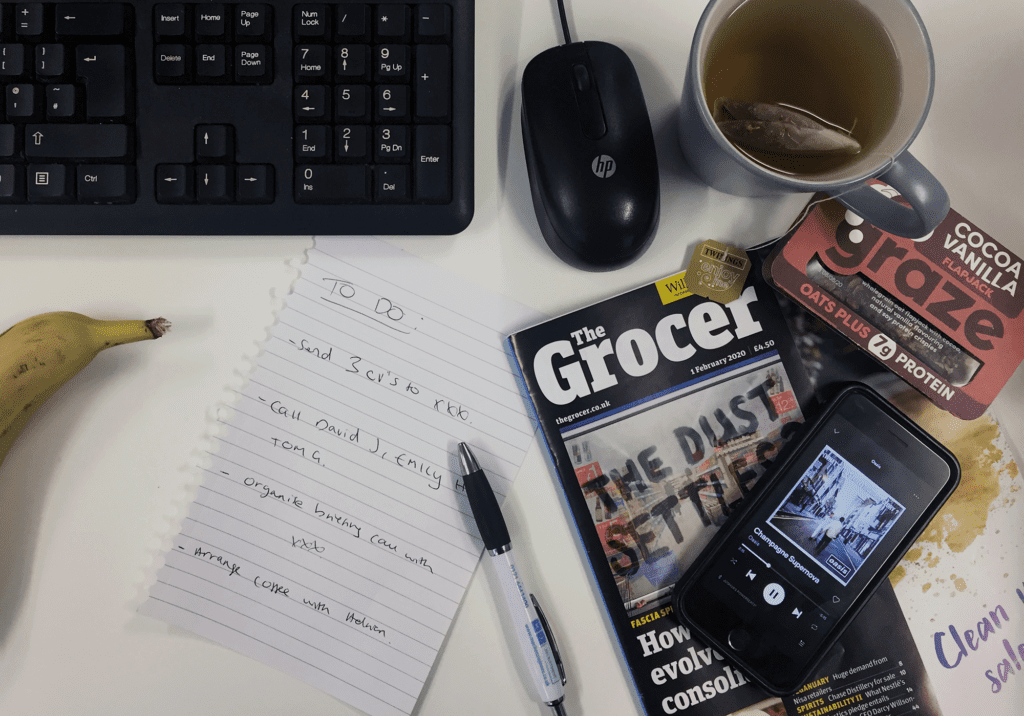 aerial shot of a desk featuring The Grocer Magazine, Twinings tea, graze protein flapjack and phone showing Oasis playing on Spotify